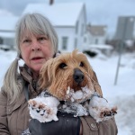 Artist photo of Char Gardner, her gray hair in pigtails. She stands in a snowy town street holding a fluffy tan dog, whose paws and muzzle are covered in snow.